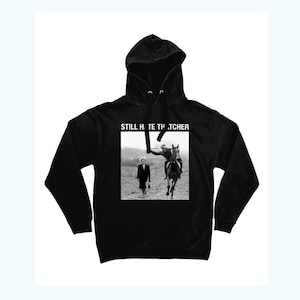 Puede incluir: Sudadera con capucha negra con un estampado gráfico blanco de una mujer caminando y un hombre a caballo. El texto "STILL HATE THATCHER" está impreso sobre el gráfico.