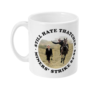 Puede incluir: Taza de cerámica blanca con un gráfico circular negro. El gráfico presenta una foto de Margaret Thatcher caminando y un hombre a caballo. El texto alrededor de la foto dice "Miners' Strike 84-85. Still Hate Thatcher."