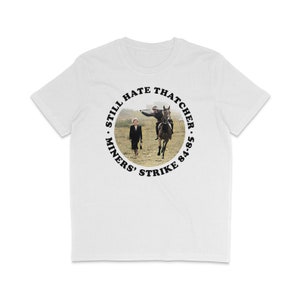 Puede incluir: Camiseta blanca con un gráfico circular negro. El gráfico presenta una foto de Margaret Thatcher montando a caballo, con una mujer caminando detrás de ella. El texto alrededor de la foto dice "Still Hate Thatcher - Miners' Strike 84-85."