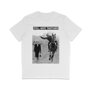 Puede incluir: Una camiseta blanca con una imagen en blanco y negro de una mujer que se aleja de un hombre a caballo. El texto "STILL HATE THATCHER" está impreso en la camiseta.