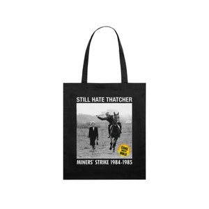 Puede incluir: Bolsa de tela negra con un gráfico blanco de una mujer que se aleja de un hombre a caballo. El gráfico incluye el texto "Still Hate Thatcher", "Miners' Strike 1984-1985", y un círculo amarillo con el texto "Coal Not Dole".