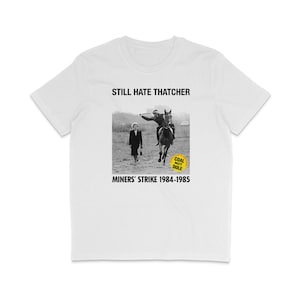 Puede incluir: Camiseta blanca con una foto en blanco y negro de una mujer que se aleja de un hombre a caballo. El texto "STILL HATE THATCHER" está en la parte superior de la imagen. El texto "MINERS' STRIKE 1984-1985" está en la parte inferior de la imagen. Un círculo amarillo con el texto "COAL NOT DOLE" está en la esquina inferior derecha de la foto.