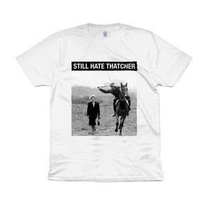 Puede incluir: Camiseta blanca con una imagen en blanco y negro de una mujer que se aleja de un hombre a caballo. El texto "STILL HATE THATCHER" está impreso en negro en la parte superior de la imagen.