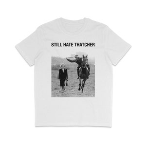 Puede incluir: Una camiseta blanca con una impresión negra que dice "STILL HATE THATCHER". La impresión también presenta una foto en blanco y negro de una mujer que se aleja de un hombre a caballo.