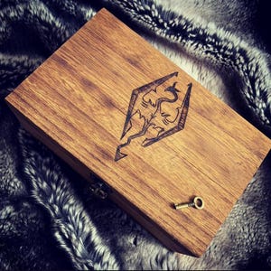 May include: A wooden box with a dragon emblem, and a small key. The box is rectangular, with a hinged lid and a brass clasp. The dragon emblem is etched into the wood. The key is small and gold-colored.