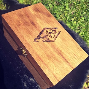 May include: A rectangular wooden box with a hinged lid and a decorative clasp. The box is made of light brown wood and features a detailed, burnt-in design on the top. The box is resting on a dark surface with green grass in the background.