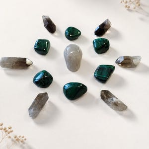 May include: An arrangement of crystals and stones, including dark green malachite and smoky quartz points, encircling a central, smooth, gray stone. The crystals vary in shape and size, creating a visually appealing display. The composition is set against a white background.