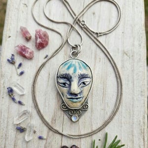 May include: A silver necklace with a pendant featuring a serene face with blue and white details. The pendant is surrounded by a silver frame and a small, round, blue stone. The necklace is displayed with crystals, lavender, and other natural elements.