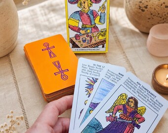 Può includere: Una scatola "STARTER TAROT DECK" con un'illustrazione colorata di una persona con un cappello e una pila di carte con il dorso arancione. Diverse carte dei tarocchi sono tenute in mano, mostrando opere d'arte e testo dettagliati. Una candela e vasi decorativi sono sullo sfondo.