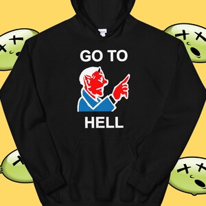 以下が含まれることがあります： 白い文字で「GO TO HELL」と書かれた黒いフード付きスウェットシャツ。赤い角と青いシャツを着た悪魔の漫画が右を指している。