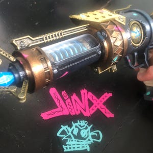 May include: A prop gun with a futuristic design, featuring bronze, gold, and purple accents. The gun has a clear tube with a blue coil inside and a blue light emanating from the barrel. The word "JINX" is displayed in pink and teal.