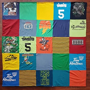 Peut inclure: Une courtepointe patchwork composée de divers t-shirts colorés. La courtepointe présente des carrés avec des motifs graphiques, du texte et des logos, dont "BEST 390 EVER", "I LIKE IKE", "TIMBITS 5" et "DUDE". La courtepointe est posée sur une surface en bois.