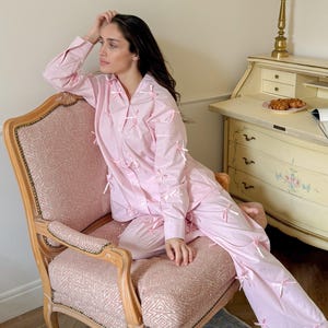 May include: Pale pink pyjama set with white trim and bow details. The long-sleeved top and trousers are worn by a person seated in a vintage pink upholstered chair. The room includes a vintage desk, decorative plates, and candle holders.