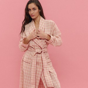 Puede incluir: Bata larga de cuadros rosa claro con ribete rojo oscuro y cinturón a juego. La bata tiene mangas largas y un diseño envolvente. El fondo es de color rosa liso.