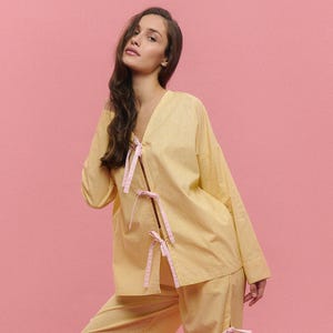 May include: Yellow gingham pajama set with pink ribbon ties. The long-sleeved top has a V-neck and the pants are wide-legged. The outfit is made of a lightweight fabric, perfect for lounging or sleepwear.