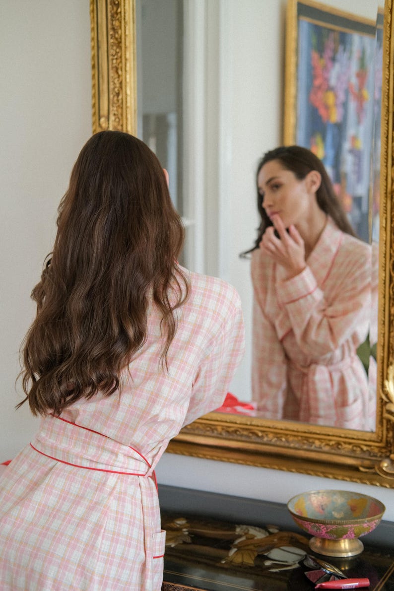 May include: A woman in a pink and white checked dressing gown with red trim stands before a gold framed mirror. The mirror reflects the woman's face and the gown. A decorative bowl sits on a table below the mirror.