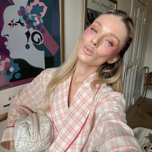 May include: A person wearing a pink and white plaid robe with red trim, holding a small floral patterned cosmetic bag. The person has pink eye patches on and is posing in front of artwork.