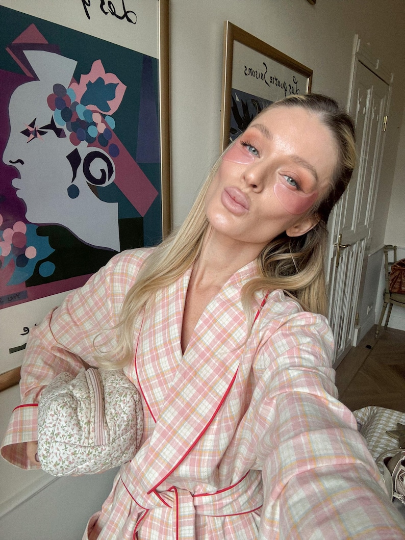 May include: A person wearing a pink and white plaid robe with red trim, holding a small floral patterned cosmetic bag. The person has pink eye patches on and is posing in front of artwork.