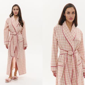 May include: A long, pink and cream plaid robe with red trim. The robe features a matching belt and pockets. The model is wearing matching pink bow slippers. The robe is designed for comfort and relaxation.