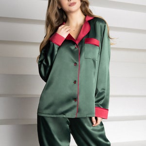 Op de afbeelding: Een groene satijnen pyjamaset met rode piping. Het bovenstuk heeft een kraag en lange mouwen. De broek heeft een rechte pijp.
