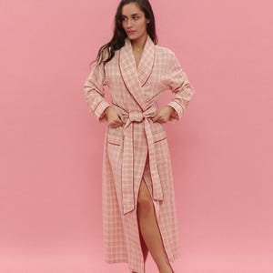 May include: A long, pale pink plaid dressing gown with red trim and a matching belt. The gown has pockets and a shawl collar. The model is wearing the gown and standing in front of a pink background.