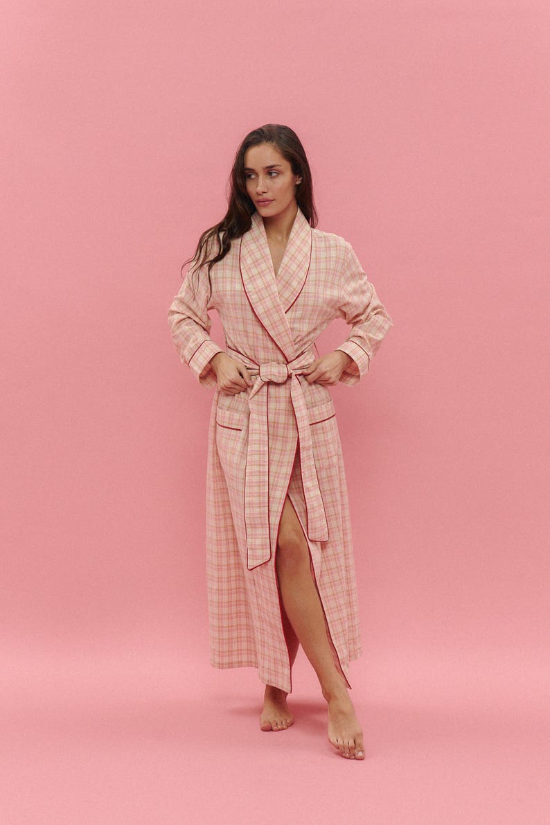 May include: A long, pale pink plaid dressing gown with red trim and a matching belt. The gown has pockets and a shawl collar. The model is wearing the gown and standing in front of a pink background.