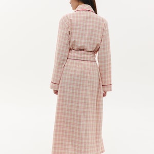 May include: A long, plaid robe in shades of pink and cream, trimmed with a deep red. The robe is belted at the waist and worn with matching pink slippers. The fabric has a classic check pattern, suitable for sleepwear or lounging.