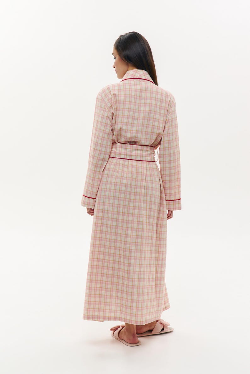 May include: A long, plaid robe in shades of pink and cream, trimmed with a deep red. The robe is belted at the waist and worn with matching pink slippers. The fabric has a classic check pattern, suitable for sleepwear or lounging.