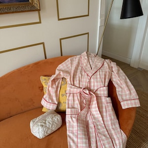 May include: A pink and white plaid robe with red trim and a matching belt is draped over an orange sofa. A small, quilted cosmetic bag and a yellow pillow with a pattern are also on the sofa. The robe has long sleeves and pockets.