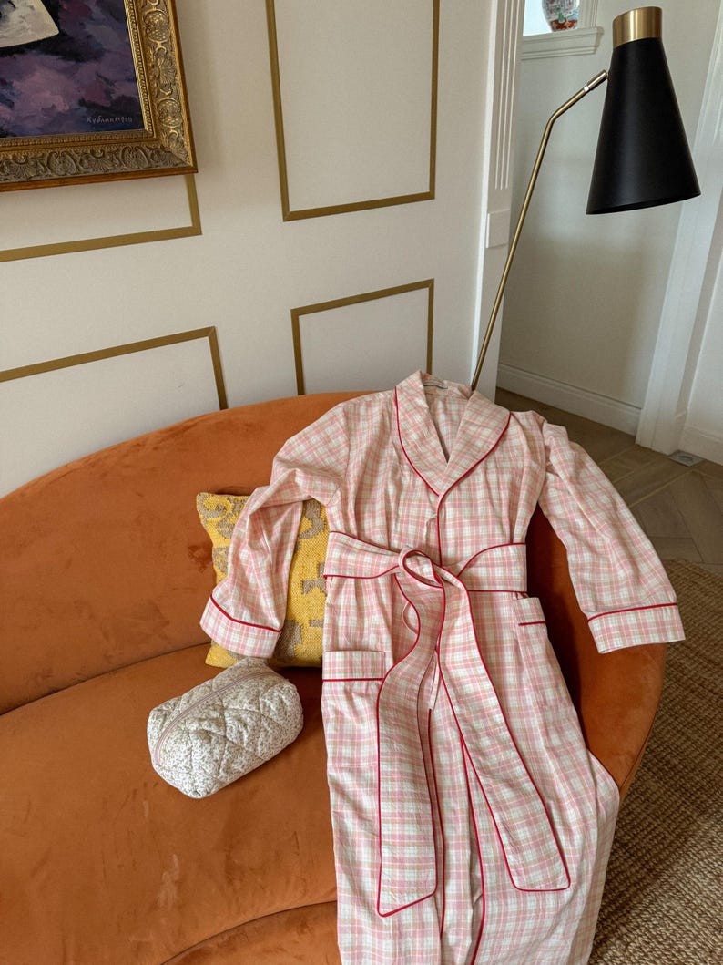 May include: A pink and white plaid robe with red trim and a matching belt is draped over an orange sofa. A small, quilted cosmetic bag and a yellow pillow with a pattern are also on the sofa. The robe has long sleeves and pockets.