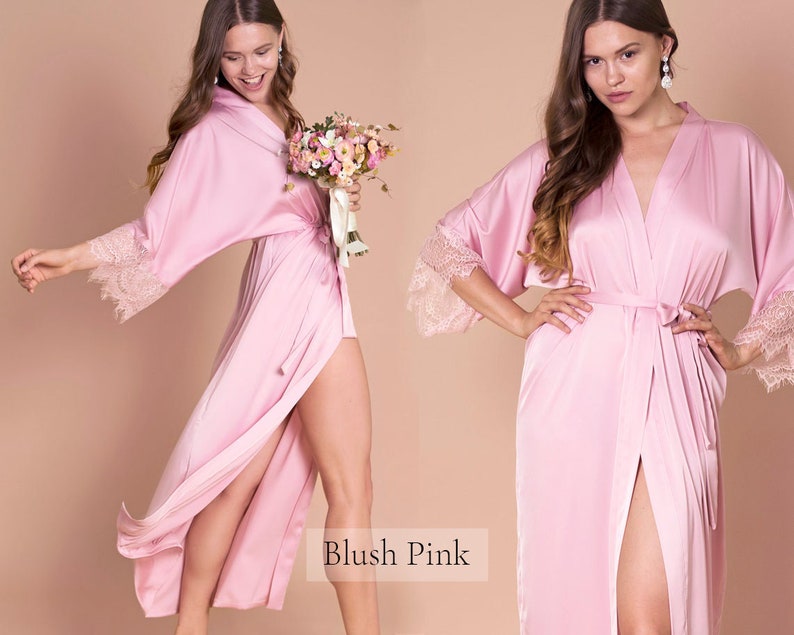 May include: A blush pink satin robe with lace trim on the sleeves. The robe has a long, flowing silhouette and a tie belt at the waist. The model is holding a bouquet of flowers.