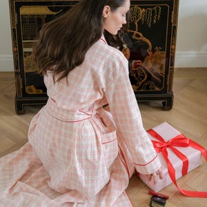 May include: A light pink plaid dressing gown with red edging and a matching belt. A wrapped gift with a red ribbon sits on the floor next to a small makeup compact. The gown is worn by a person kneeling on a wooden floor.