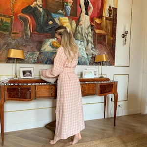 May include: A woman wearing a pink and white plaid robe stands in front of a wooden desk with drawers. A large painting with a red background hangs on the wall behind her. Two lamps with gold shades sit on the desk.