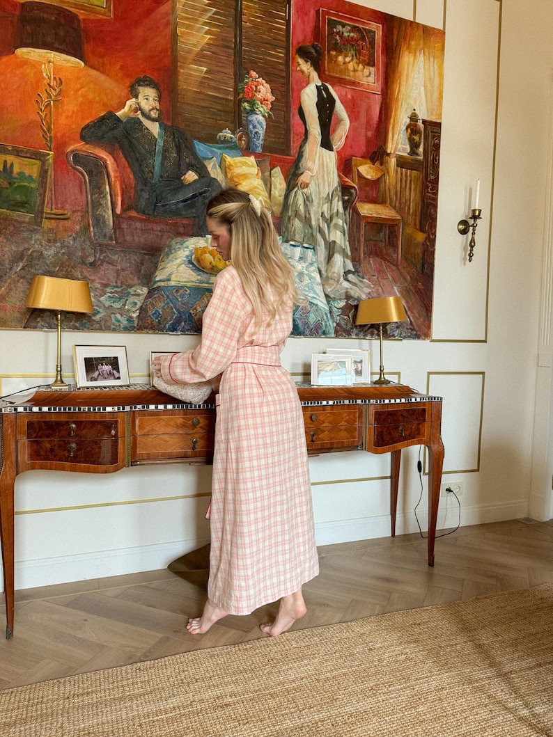 May include: A woman wearing a pink and white plaid robe stands in front of a wooden desk with drawers. A large painting with a red background hangs on the wall behind her. Two lamps with gold shades sit on the desk.