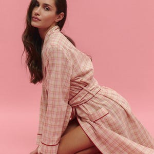 May include: A light pink plaid robe with red trim. The robe is made of a soft fabric and has long sleeves. The model is kneeling on a pink background. The robe is tied at the waist.