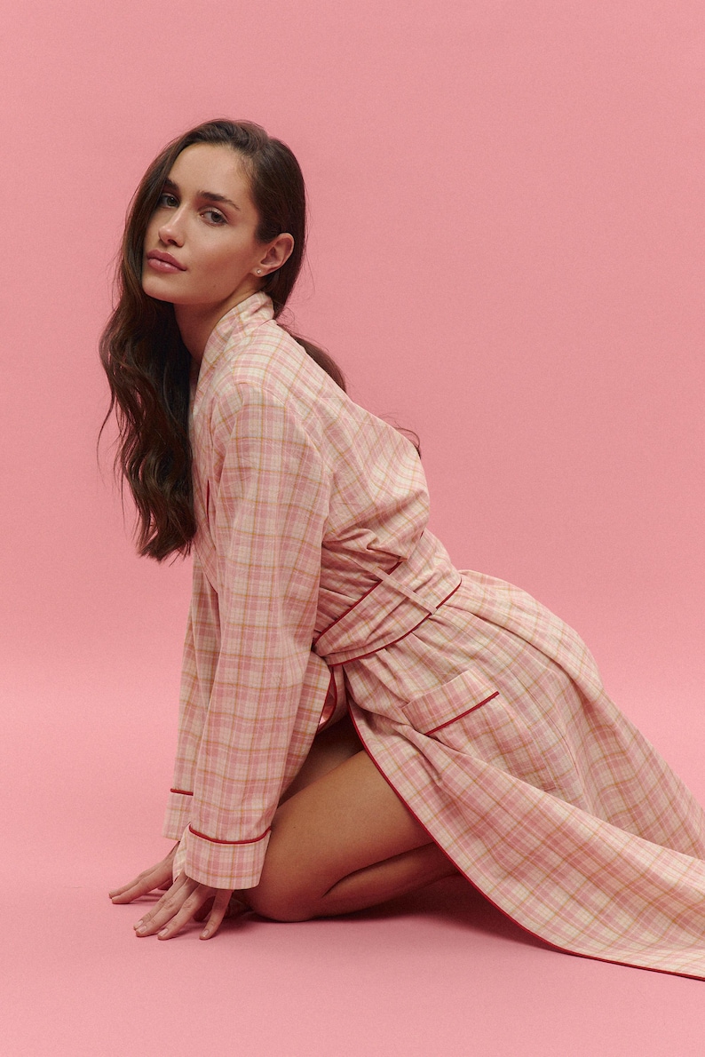 May include: A light pink plaid robe with red trim. The robe is made of a soft fabric and has long sleeves. The model is kneeling on a pink background. The robe is tied at the waist.