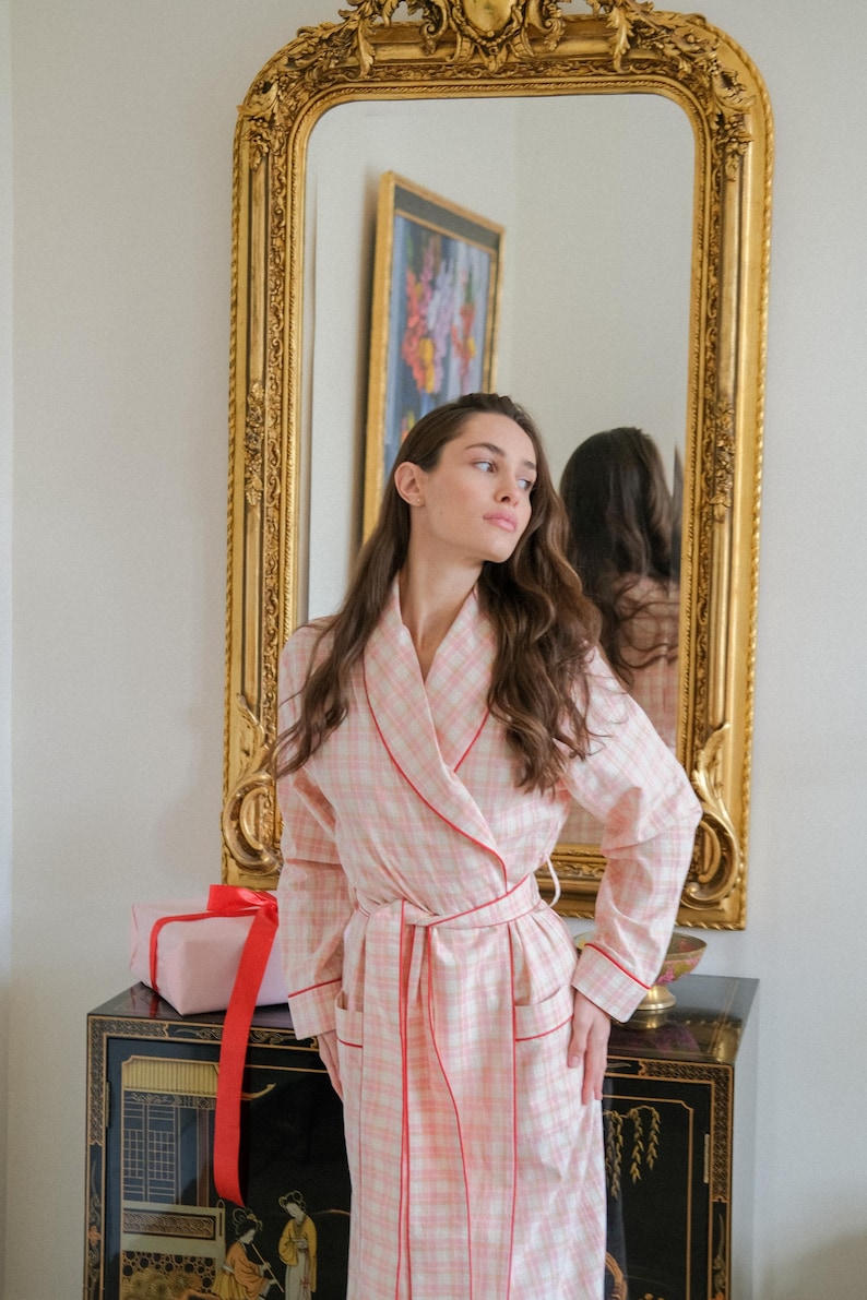 May include: A pink plaid dressing gown with red trim and a matching belt. The gown is worn over clothing. A pink gift box with a red ribbon sits on a black cabinet. A gold framed mirror is behind the gown.