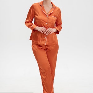 Op de afbeelding: Satijnen pyjamaset in verbrand oranje met witte piping. Het bovenstuk heeft een knoopsluiting en een borstzak. De broek heeft een rechte pijp.