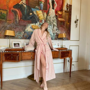 May include: A pink and white plaid robe with red trim. The robe is open, revealing the legs. The background features a large painting, a wooden desk with lamps, and a woven rug.
