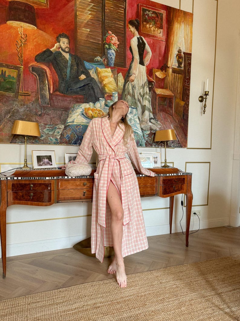 May include: A pink and white plaid robe with red trim. The robe is open, revealing the legs. The background features a large painting, a wooden desk with lamps, and a woven rug.