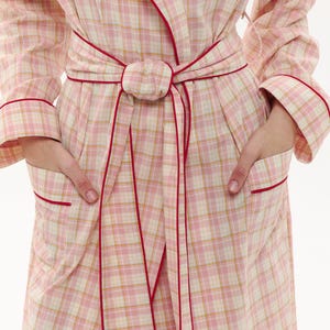May include: A pink plaid dressing gown with red trim and a matching belt. The gown features long sleeves, pockets, and a wrap-around design. The plaid pattern combines pink, white, and yellow hues, creating a soft aesthetic. Designed for comfort and relaxation, it's a cosy garment.