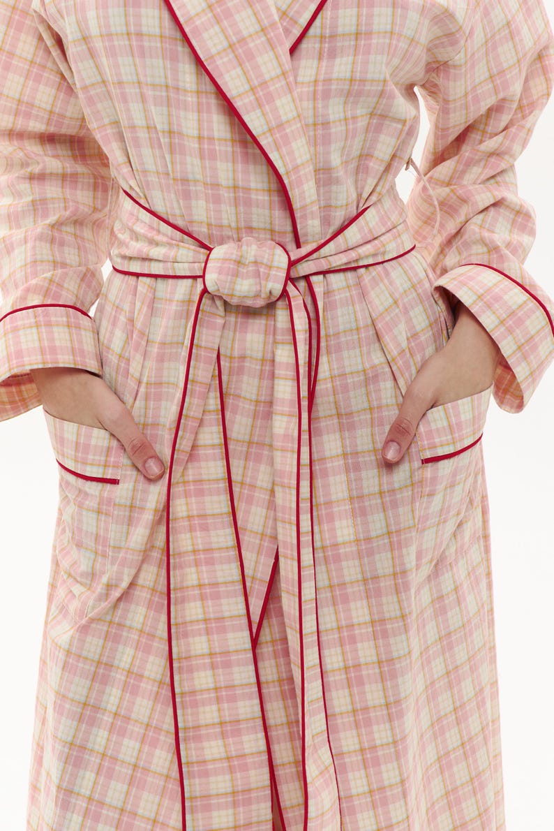 May include: A pink plaid dressing gown with red trim and a matching belt. The gown features long sleeves, pockets, and a wrap-around design. The plaid pattern combines pink, white, and yellow hues, creating a soft aesthetic. Designed for comfort and relaxation, it's a cosy garment.