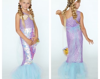 May include: A young girl wearing a purple and iridescent mermaid costume with a white crown. The costume features a sequined bodice with a seashell design and a tulle skirt.