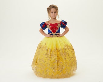 May include: A young girl dressed as Snow White in a yellow and gold dress with a blue and red bodice. She is standing with her hands on her hips and looking at the camera.