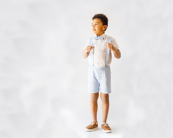 May include: A young boy wearing a white button-down shirt with a light blue bow tie and matching light blue shorts. He is wearing brown shoes and has suspenders.