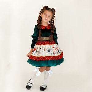 Puede incluir: Una niña pequeña con un vestido de terciopelo verde con una falda roja y blanca que representa una escena navideña. El vestido tiene un cinturón dorado y un cuello blanco estilo Peter Pan. Lleva calcetines blancos con lazos y zapatos negros.