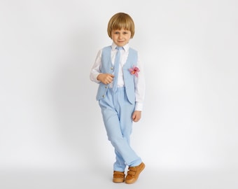 May include: A young boy wearing a light blue suit with a white shirt and a light blue tie. He is wearing a pink flower on his lapel.