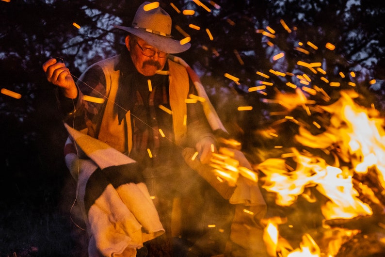 May include: A man wearing a brown hat and a brown vest sits by a campfire. He is holding a stick in his hand and there are sparks flying from the fire.