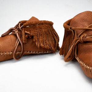 May include: A pair of brown leather moccasins with fringe and laces. The moccasins are handmade and have a traditional design.