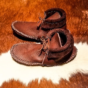 May include: A pair of brown leather moccasins with suede trim. The moccasins are on a white and brown cowhide rug.